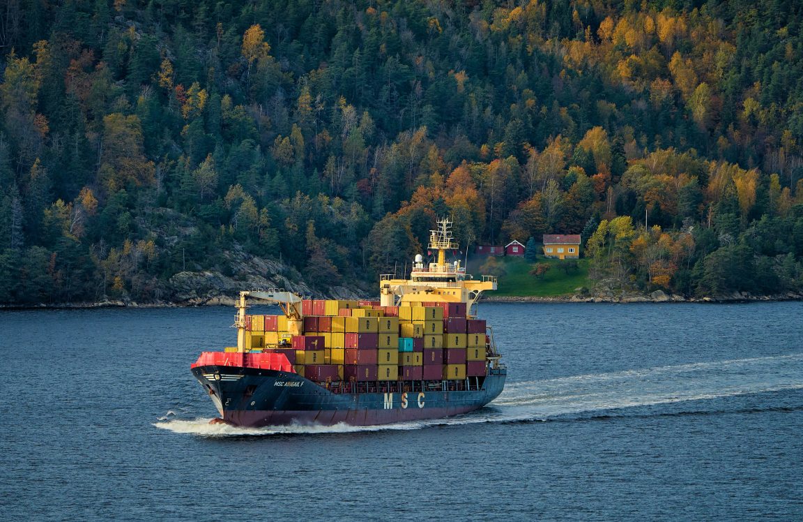 Container ship loaded with stacked cargo containers sails across a calm bay with forested hills and a few houses in the background.